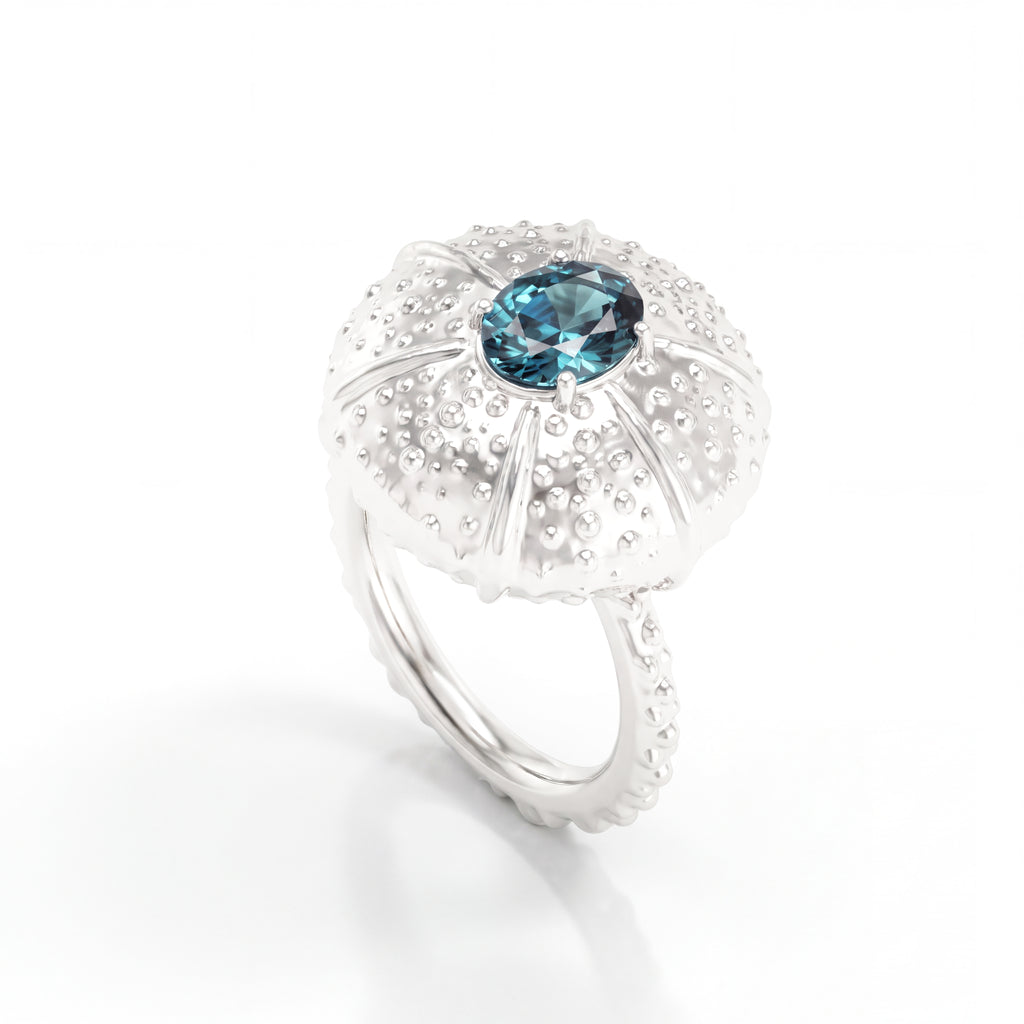 A white gold ring featuring a textured, sea urchin-inspired dome setting with a central oval teal saphire, shown on a clean white background.