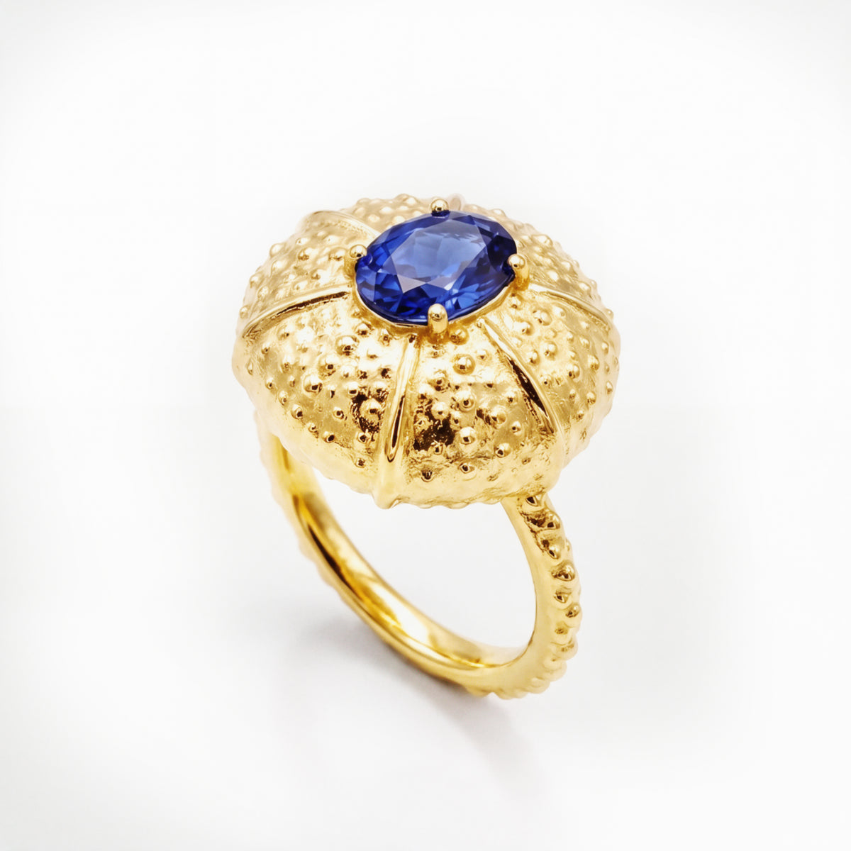 A yellow gold ring featuring a textured, sea urchin-inspired dome setting with a central blue sapphire, shown on a clean white background.