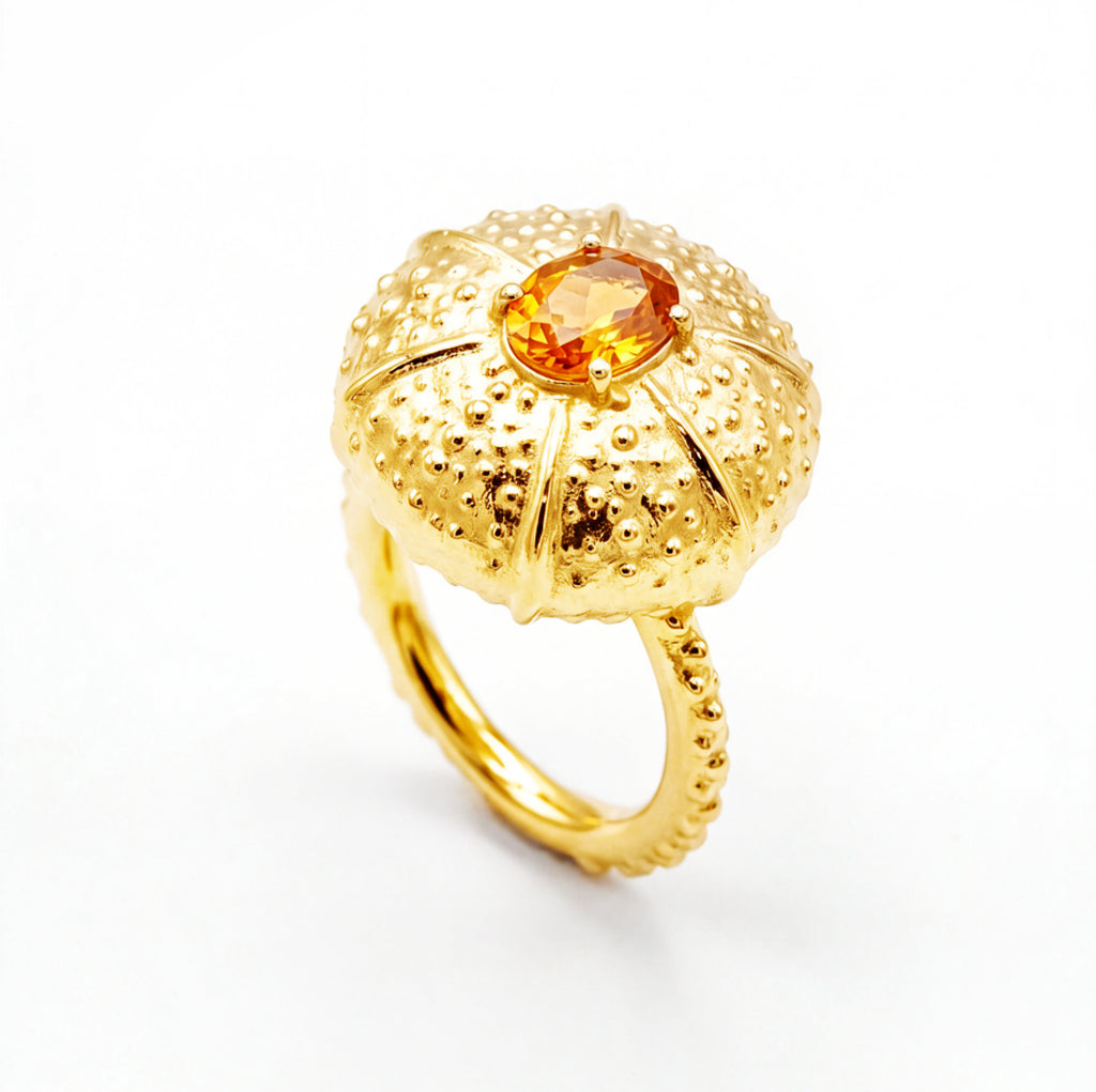 A gold ring designed like a sea urchin featuring a textured dome with a central faceted orange sapphire, set against a clean white background.