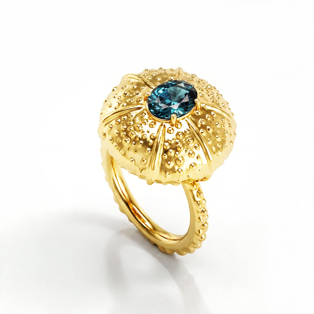 A yellow gold ring featuring a textured, sea urchin-inspired dome setting with a central oval teal sapphire, shown on a clean white background.