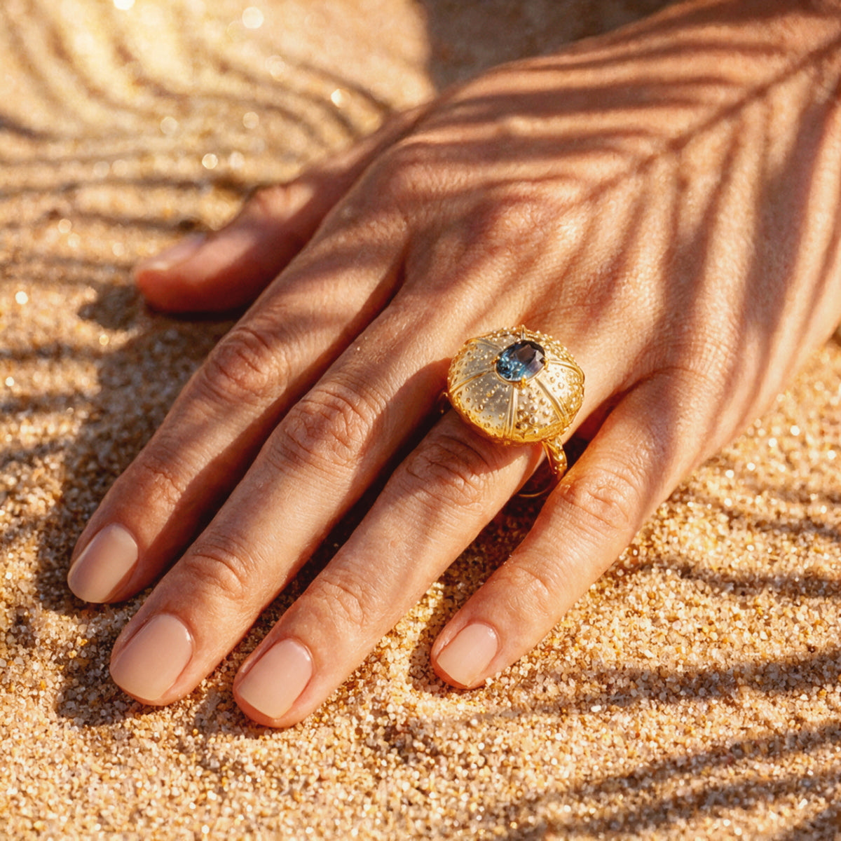 A yellow gold ring featuring a textured, sea urchin-inspired dome setting with a central teal sapphire, worn on a hand resting on sandy ground under leaf shadows.