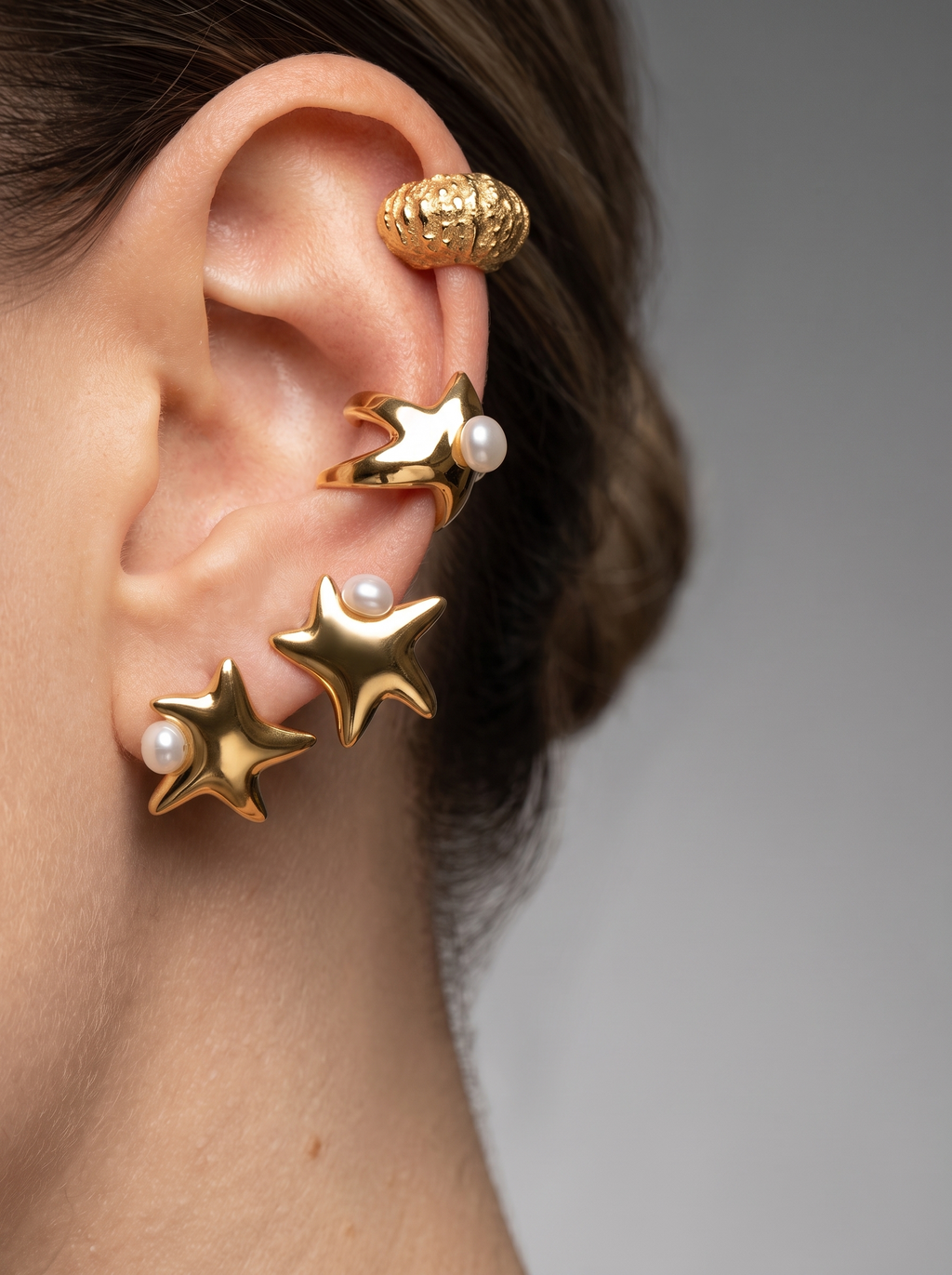 A close-up side profile of a person's ear wearing multiple gold-toned star-shaped earrings, including two with small white pearl accents and one textured gold ear cuff.